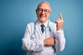 Senior grey haired doctor man wearing stethoscope and medical coat over blue background with a big smile on face, pointing with Royalty Free Stock Photo