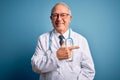 Senior grey haired doctor man wearing stethoscope and medical coat over blue background cheerful with a smile of face pointing Royalty Free Stock Photo