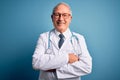 Senior grey haired doctor man wearing stethoscope and medical coat over blue background happy face smiling with crossed arms Royalty Free Stock Photo