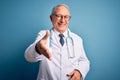 Senior grey haired doctor man wearing stethoscope and medical coat over blue background smiling friendly offering handshake as Royalty Free Stock Photo