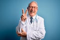 Senior grey haired doctor man wearing stethoscope and medical coat over blue background smiling with happy face winking at the Royalty Free Stock Photo