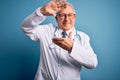 Senior grey haired doctor man wearing stethoscope and medical coat over blue background smiling making frame with hands and Royalty Free Stock Photo