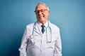 Senior grey haired doctor man wearing stethoscope and medical coat over blue background winking looking at the camera with sexy Royalty Free Stock Photo