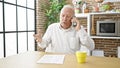 Senior grey-haired man angry arguing on smartphone sitting on table at dinning room