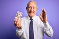 Senior grey haired man holding bunch of fifty dollars banknotes over purple background doing ok sign with fingers, excellent