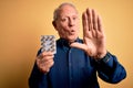 Senior grey haired man holding pharmaceutical pill drugs over yellow background with open hand doing stop sign with serious and