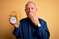 Senior grey haired man holding vintage alarm clock over yellow background cover mouth with hand shocked with shame for mistake, Royalty Free Stock Photo