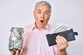 Senior grey-haired man holding wallet with dollars and jar with savings afraid and shocked with surprise and amazed expression, Royalty Free Stock Photo