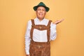 Senior grey-haired man wearing german traditional octoberfest suit over yellow background smiling cheerful presenting and pointing