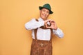 Senior grey-haired man wearing german traditional octoberfest suit over yellow background smiling in love doing heart symbol shape