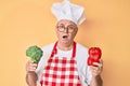 Senior grey-haired man wearing professional cook apron holding broccoli and red pepper in shock face, looking skeptical and Royalty Free Stock Photo