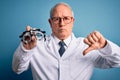 Senior grey haired optic doctor man holding optometrist eyeglasses over blue background with angry face, negative sign showing Royalty Free Stock Photo