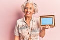 Senior grey-haired woman holding empty frame looking positive and happy standing and smiling with a confident smile showing teeth Royalty Free Stock Photo