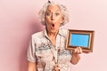 Senior grey-haired woman holding empty frame scared and amazed with open mouth for surprise, disbelief face Royalty Free Stock Photo