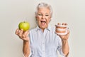 Senior grey-haired woman holding green apple and denture teeth smiling and laughing hard out loud because funny crazy joke Royalty Free Stock Photo