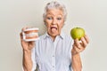 Senior grey-haired woman holding green apple and denture teeth sticking tongue out happy with funny expression