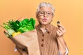 Senior grey-haired woman holding groceries and 1 euro coin puffing cheeks with funny face