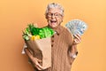 Senior grey-haired woman holding groceries and united states dollars banknotes smiling and laughing hard out loud because funny