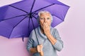 Senior grey-haired woman holding purple umbrella covering mouth with hand, shocked and afraid for mistake