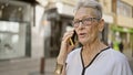 Senior grey-haired woman masters modern technology, engrossed in serious phone conversation on sunny city street Royalty Free Stock Photo