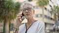 Senior grey-haired woman masters modern technology, engrossed in serious phone conversation on sunny city street Royalty Free Stock Photo