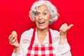 Senior grey-haired woman wearing apron holding egg pointing thumb up to the side smiling happy with open mouth Royalty Free Stock Photo