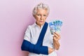 Senior grey-haired woman wearing arm on sling holding south africa rands banknotes depressed and worry for distress, crying angry