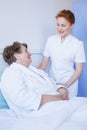 Senior woman lying in white hospital bed with young helpful nurse holding her hand Royalty Free Stock Photo