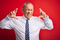 Senior handsome businessman wearing elegant tie standing over isolated red background gesturing finger crossed smiling with hope