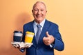 Senior handsome grey-haired businessman wearing suit holding cups of takeaway coffee smiling happy and positive, thumb up doing Royalty Free Stock Photo