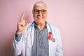 Senior handsome hoary doctor man wearing stethoscope and red HIV ribbon on coat pointing finger up with successful idea