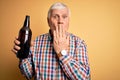 Senior handsome hoary man drinking bottle of beer standing over isolated yellow background cover mouth with hand shocked with