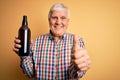 Senior handsome hoary man drinking bottle of beer standing over isolated yellow background happy with big smile doing ok sign,