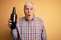 Senior handsome hoary man drinking bottle of beer standing over isolated yellow background scared in shock with a surprise face,