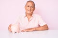 Senior handsome man with gray hair holding piggy bank sitting on the table looking positive and happy standing and smiling with a Royalty Free Stock Photo