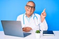 Senior handsome man with gray hair wearing doctor uniform working using computer laptop with a big smile on face, pointing with Royalty Free Stock Photo