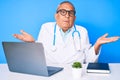 Senior handsome man with gray hair wearing doctor uniform working using computer laptop clueless and confused expression with arms Royalty Free Stock Photo