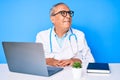 Senior handsome man with gray hair wearing doctor uniform working using computer laptop looking away to side with smile on face, Royalty Free Stock Photo