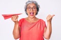 Senior hispanic grey- haired woman holding paper airplane pointing thumb up to the side smiling happy with open mouth Royalty Free Stock Photo