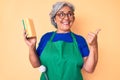 Senior hispanic grey- haired woman wearing apron holding scourer pointing thumb up to the side smiling happy with open mouth Royalty Free Stock Photo