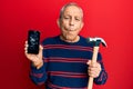 Senior hispanic man holding broken smartphone showing cracked screen and hammer making fish face with mouth and squinting eyes,