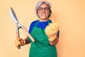 Senior hispanic woman wearing gardener apron and gloves holding shears annoyed and frustrated shouting with anger, yelling crazy