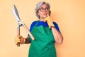 Senior hispanic woman wearing gardener apron and gloves holding shears serious face thinking about question with hand on chin,