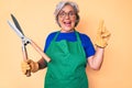 Senior hispanic woman wearing gardener apron and gloves holding shears surprised with an idea or question pointing finger with