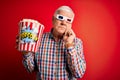 Senior hoary man watching film using 3d glasses eating popcorn over red background serious face thinking about question, very