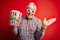 Senior hoary man watching film using 3d glasses eating popcorn over red background very happy and excited, winner expression