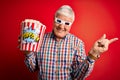 Senior hoary man watching film using 3d glasses eating popcorn over red background very happy pointing with hand and finger to the