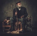 Senior hunter with a english setter and shotgun in a traditional shooting clothing, sitting on a dark background.