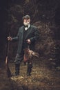 Senior hunter with a shotgun and pheasants in a traditional shooting clothing, posing on a dark forest background.