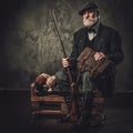 Senior hunter with a shotgun and pheasants in a traditional shooting clothing, sitting on a dark background. Royalty Free Stock Photo